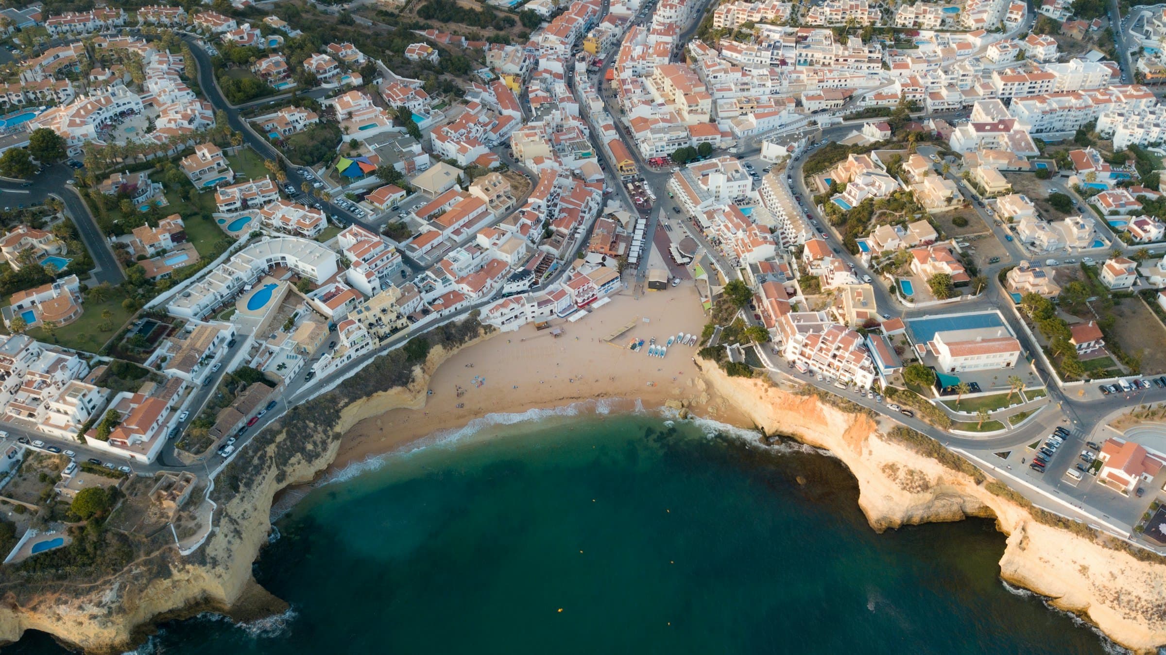 Vista aérea de uma vila costeira do Algarve: centenas de moradias, apartamentos e hotéis em redor de uma praia entre falésias.